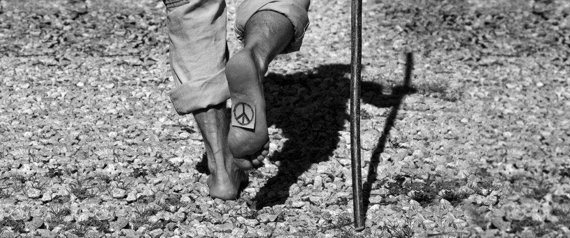 Mann barfuß unterwegs auf Steinweg mit Pilgerstock und Friedenszeichen auf der Fußsohle