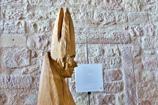Holzskulptur eines Bischofs mit Mitra vor einer groben Sandsteinwand, daneben ein beschriftetes Schild.