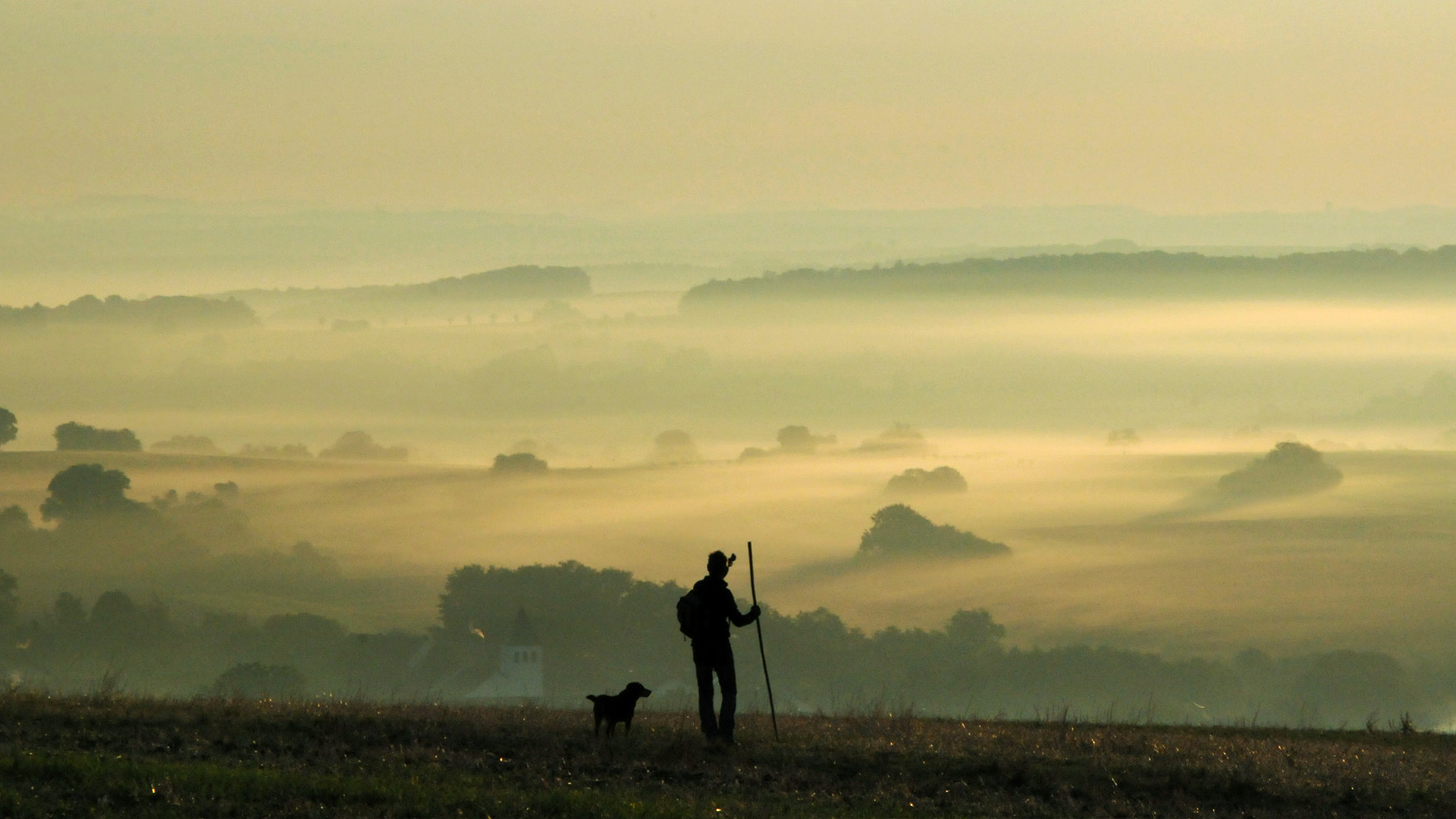 Pilger mit Hund vor Panorama einer nebelverhangenen Herbstlandschaft